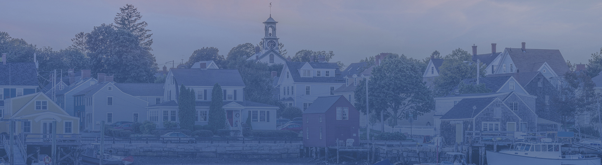 Panoramic view of a historic coastal town with tree-lined homes and a clock-tower church beneath an overcast sky.