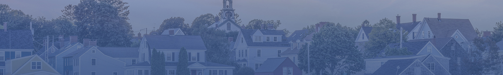Panoramic view of a historic coastal town with tree-lined homes and a clock-tower church beneath an overcast sky.